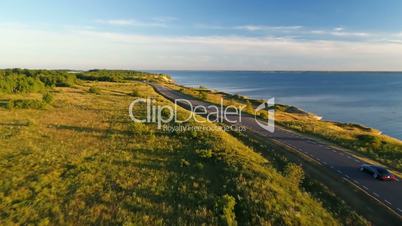 Aerial view of driving black car on the road along the cliff near the sea in summer at sunset