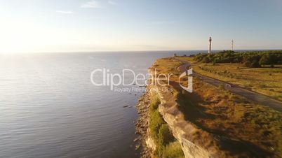 Aerial view of driving black car along the sea cliff in summer at sunset. In the background the old lighthouse.