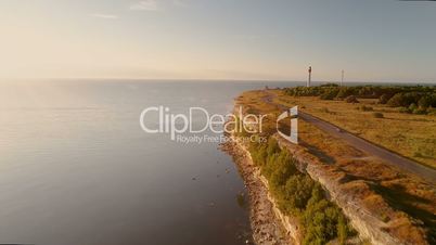 Aerial view of driving black car along the sea cliff in summer at sunset. In the background the old lighthouse.