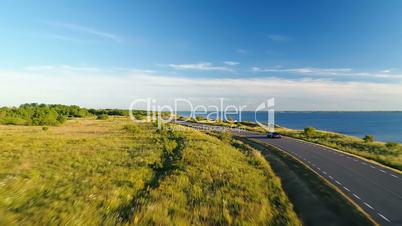 Aerial view of the driving black car on the road along the cliff next the sea in summer at sunset