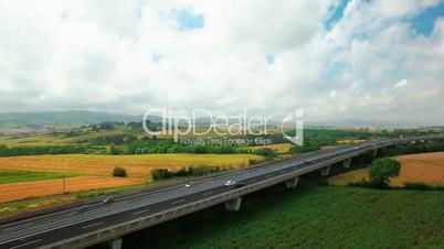 Aerial view of driving vehicles by the highway between yellow and green grass. Aerial view of fields and rural settlements by summer sunny day in Tuscany, Italy
