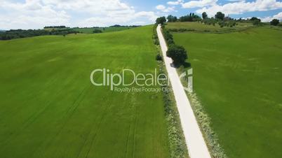 Aerial flight over the field along the rural road with driving white color car. In the Background of blue sky and clouds. Italy, Tuscany