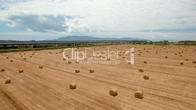 Aerial flight over the fields and next the highway. Aerial view with background of blue sky and clouds and mountains. On the fields are stored bales of hay next the highway