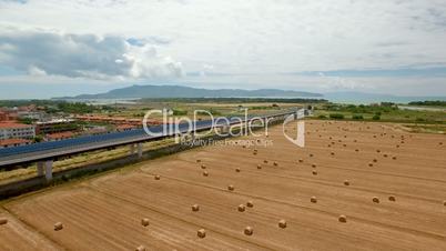 Aerial flight over the fields and next the highway. Aerial view with background of blue sky and clouds and mountains. On the fields are stored bales of hay next the highway
