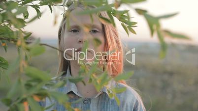 A little girl with a sad face looks through the branches of a leafy tree. Close-up. Slow motion.