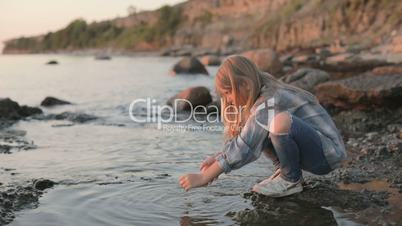 A little girl washes her hands and face with sea water by the sea coast. Sunset at the sea.