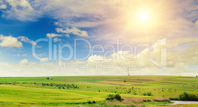 Hilly green field, windmill and sun on blue sky background. Wide