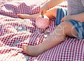Baby Boy Sitting on Picnic Blanket PUtting Coins in Piggy Bank