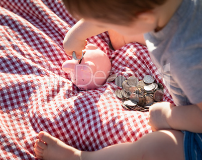 Baby Boy Sitting on Picnic Blanket PUtting Coins in Piggy Bank