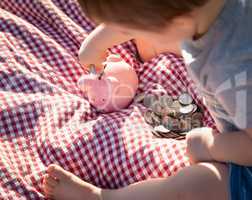 Baby Boy Sitting on Picnic Blanket PUtting Coins in Piggy Bank
