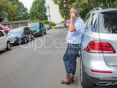 Man is standing in the street next to car and talking on the phone