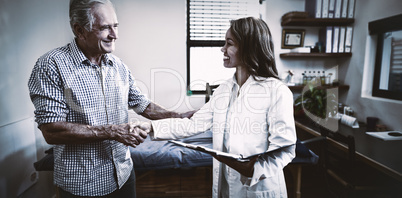 Smiling senior male patient and female therapist shaking hands against window