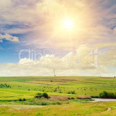 Hilly green field, windmill and sun on blue sky background.