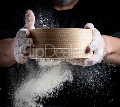man sifts white wheat flour through a wooden sieve