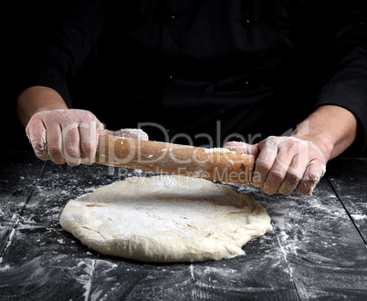 chef in a black tunic rolls a dough for a round pizza