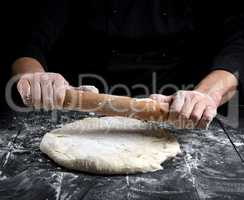 chef in a black tunic rolls a dough for a round pizza