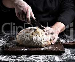 man cuts with a knife a round whole loaf of white wheat flour