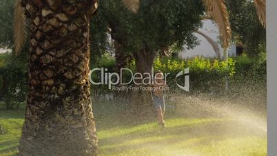 Child running on the lawn and getting wet from water sprinkler