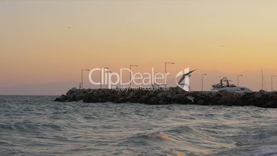 Waterscape with wavy sea, quay and flying seagulls at sunset