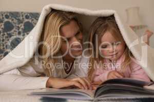 Mother with her daughter reading storybook under blanket in bedroom