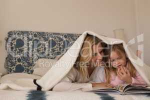 Mother with her daughter reading storybook under blanket in bedroom