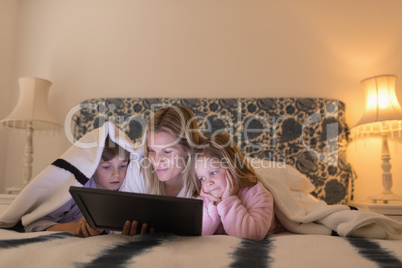 Mother with her children using digital tablet under blanket in bedroom