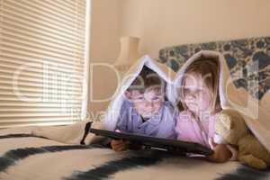 Siblings using digital tablet under blanket in bedroom