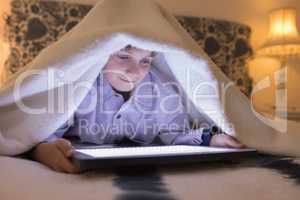 Boy under blanket using digital tablet in bedroom at home