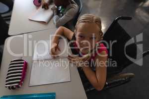 Overhead of disable schoolgirl looking at camera while sitting at desk in classroom