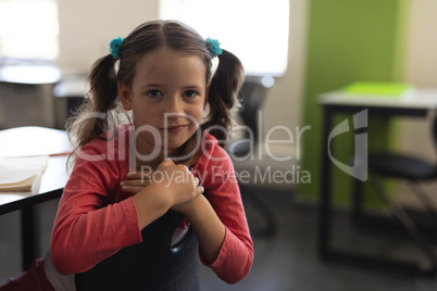 Front view of cute schoolgirl looking at camera and sitting at desk in classroom