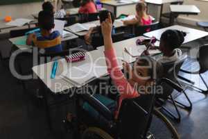 High angle view of disable schoolgirl looking at camera and raising hand in classroom