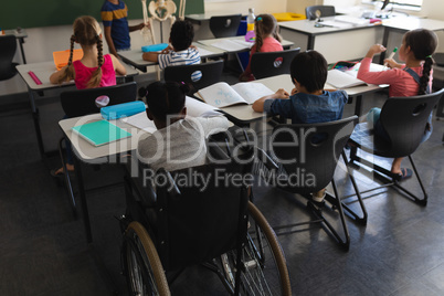 Rear view of disable schoolboy with classmate studying and sitting at desk in classroom