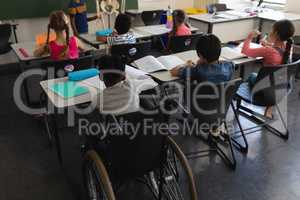 Rear view of disable schoolboy with classmate studying and sitting at desk in classroom