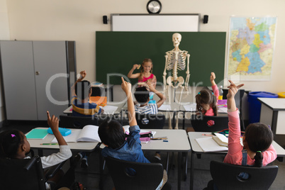 Rear view of schoolkids raising hands and sitting at desk in classroom