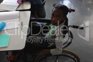 High angle view of disable black schoolgirl looking at camera and sitting at desk in classroom