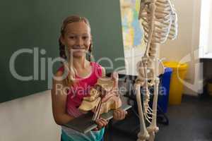 Smiling schoolgirl holding anatomical model and looking at camera in classroom