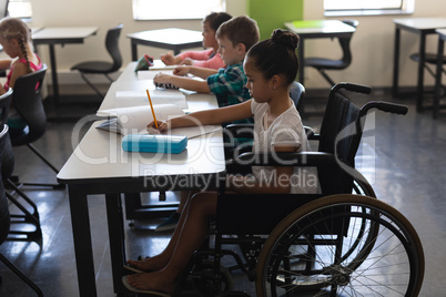 Side view of disabled schoolgirl with classmates studying and sitting at desk in classroom