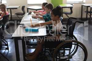 Side view of disabled schoolgirl with classmates studying and sitting at desk in classroom