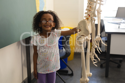 Front view of little happy black schoolgirl holding human skeleton model and looking at camera in cl