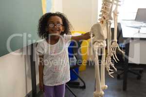 Front view of little happy black schoolgirl holding human skeleton model and looking at camera in cl