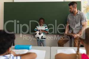 Front view of black schoolboy holding football and standing at green board in classroom