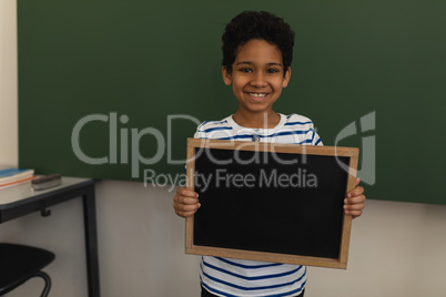 Front view of happy schoolboy holding slate board and looking at camera in classroom