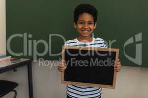 Front view of happy schoolboy holding slate board and looking at camera in classroom