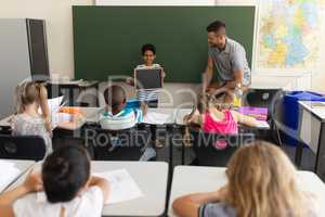 Front view of happy schoolboy holding slate board and standing at greenboard in classroom