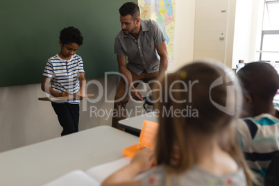 Schoolboy writing on slate board and teacher looking at him in classroom