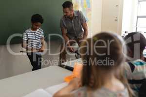 Schoolboy writing on slate board and teacher looking at him in classroom