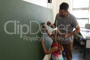Side view of schoolgirl with math teacher writing on chalk board in classroom