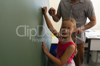 Schoolgirl with math teacher looking at camera and writing on chalk board in classroom