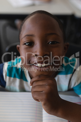Close-up of black schoolboy with hand on chin sitting at desk and looking at camera in classroom