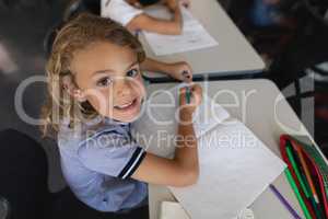 High angle view of smiling schoolgirl studying at desk and looking at camera in classroom